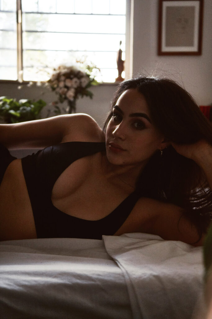 Valeria Cisneros lounges gracefully on a bed wearing a black bralette with a serene ambiance illuminated by natural light filtering through a window.